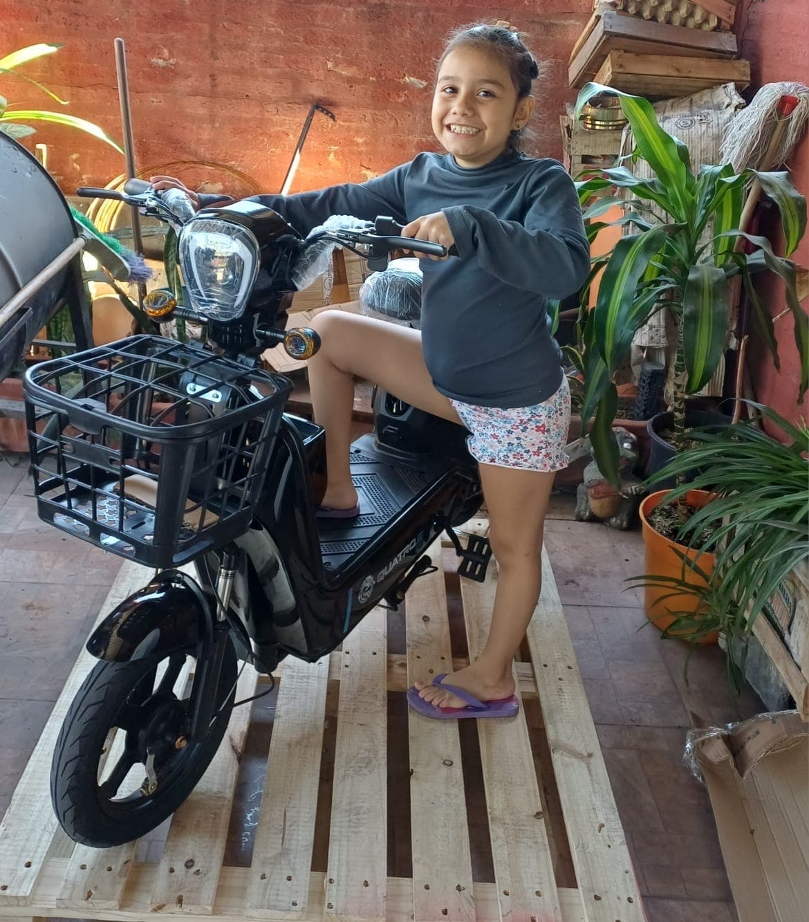 Nena sentada en la moto eléctrica nueva, sobre el pallet en que llegó